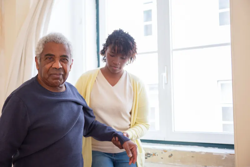 Home health aide helping a senior man walk at home beside a bright window